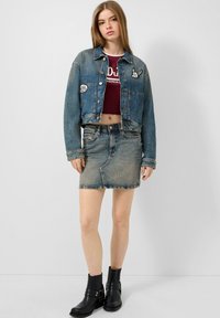 Jeansjacke mit verkürztem Schnitt, blauer Farbe, Knopfleiste und Aufnähern. Kombiniert mit einem körperbetonten burgunderfarbenen Oberteil und einem verwaschenen Jeansrock. Schwarze Ankle-Boots.