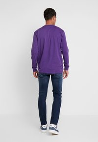 Lila långärmad tröja med rund halsringning, kombinerad med mörkblå jeans och vita sneakers, visad från bakview.