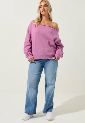 Lyshåret kvinde iført off-shoulder lavendel sweatshirt, lyseblå jeans med vide ben og hvide sneakers, stående foran en ensfarvet baggrund.