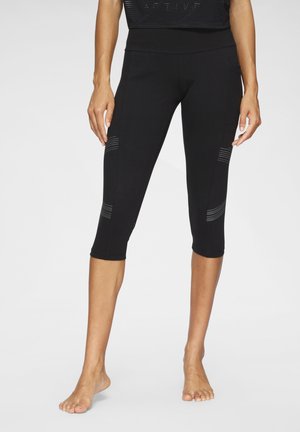 Woman standing barefoot wearing black cropped leggings with subtle horizontal stitch details on thighs and calves.