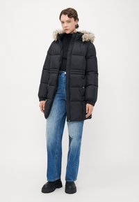 Giovane in giacca a vento nera con cappuccio in pelliccia, dolcevita nero, jeans blu e stivali neri in piedi su uno sfondo semplice.
