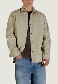 Camicia color beige chiaro con bottoni, realizzata in tessuto liscio, con una tasca sul petto, bottoni a pressione e una vestibilità comoda, indossata sopra una camicia marrone scuro.