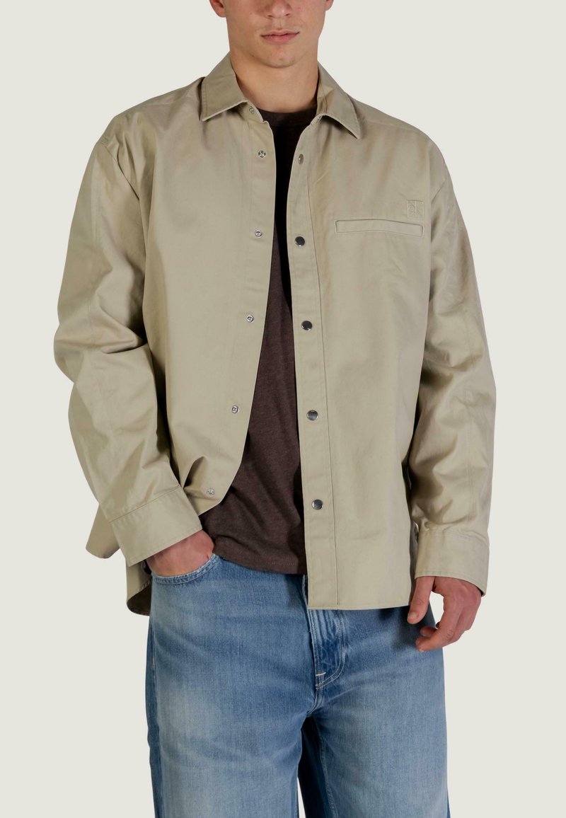 Camicia color beige chiaro con bottoni, realizzata in tessuto liscio, con una tasca sul petto, bottoni a pressione e una vestibilità comoda, indossata sopra una camicia marrone scuro.