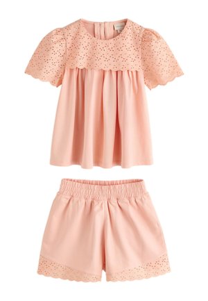 Junges Mädchen mit langen blonden Haaren, das eine pfirsichfarbene bestickte Bluse und passende Shorts trägt, steht mit einem leichten Lächeln vor einem schlichten Hintergrund.