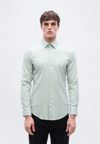 Νεαρός άνδρας με κοντά σγουρά μαλλιά, φοράει εφαρμοστό ανοιχτοπράσινο πουκάμισο με κουμπιά και μαύρο παντελόνι, στέκεται μπροστά σε ένα απλό λευκό φόντο.