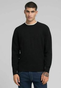 Pull noir à manches longues, col rond avec bordure blanche, confectionné dans un tissu à texture douce, porté avec un jean bleu.