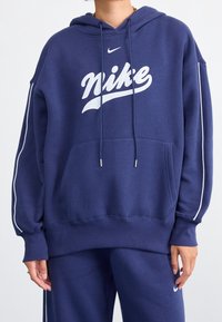 Tmavě modrá mikina s kapucí vyrobená z mikinového materiálu s velkým bílým logem Nike, šňůrkami, přední kapsou a bílými bočními pruhy.