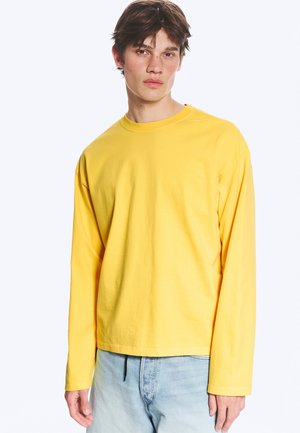 Jonge man die een felgeel shirt met lange mouwen en lichtblauwe spijkerbroek draagt, staand tegen een effen witte achtergrond.