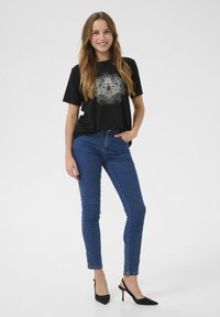 T-shirt nera con design grafico argentato, jeans skinny in denim blu e décolleté nere con zeppa. La modella sta in piedi con una mano in tasca.