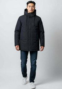 Jeune homme portant un manteau d'hiver noir avec capuche, à fermeture éclair et boutons, un jean bleu et des baskets blanches, se tenant sur un fond neutre.