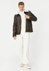 Brun faux læderjakke med hvidt shearling krave, lynlås lukning, to sidelommer; båret over en hvid sweater og lyse bukser.