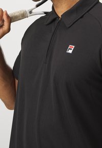 Czarny polo z zamkiem błyskawicznym i subtelną teksturą pasków, z małym, wielokolorowym logo na wysokości klatki piersiowej.