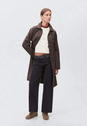 Brauner Knopfverschlussmantel über einem weißen Oberteil und schwarzen weit geschnittenen Hosen mit Nieten-Gürtel. Braune Sneakers runden das Outfit ab. Insgesamt glatte Textur.
