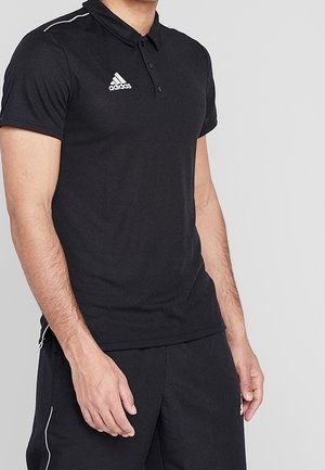 Homme portant un polo noir Adidas et un short noir avec des liserés blancs, debout devant un fond gris uni.