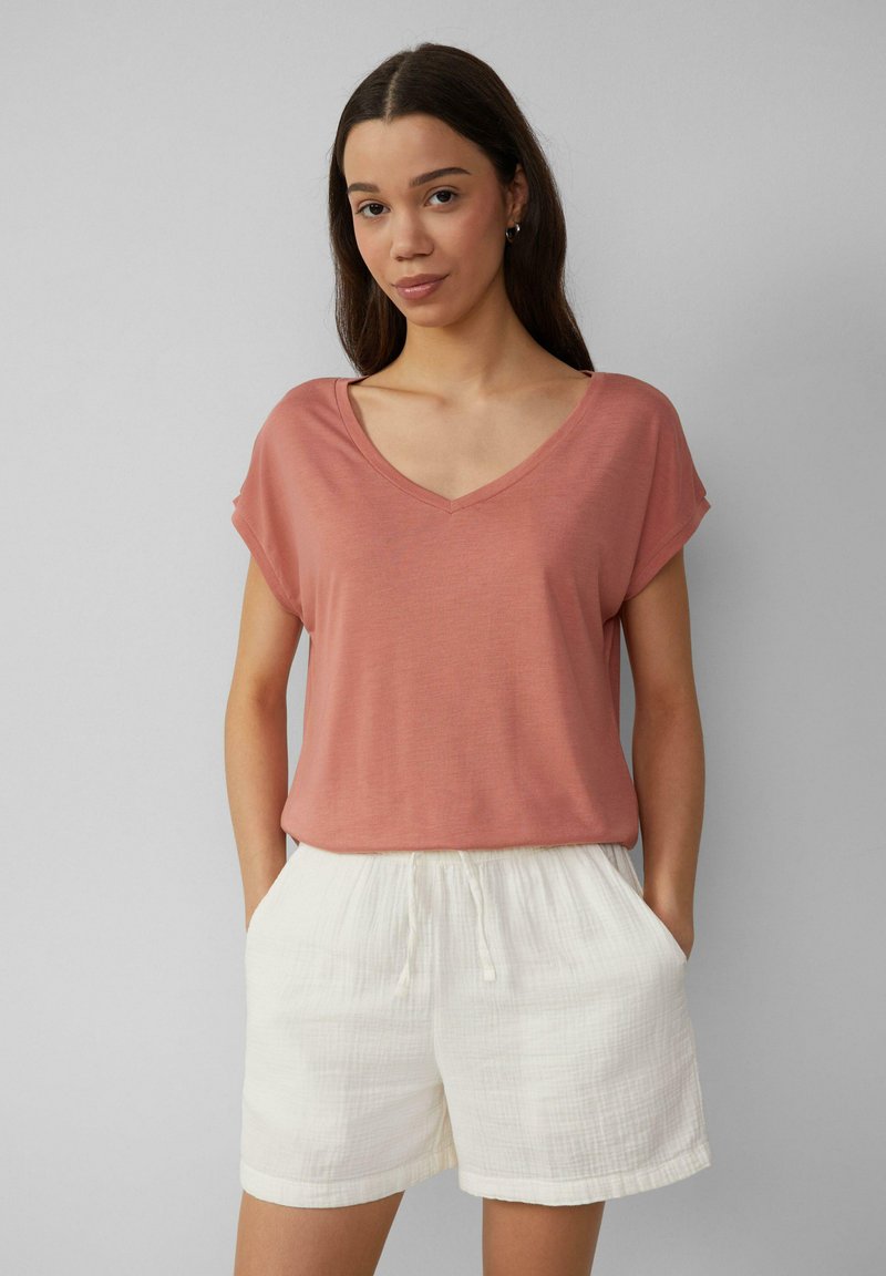 Rozā krāsas V-veida kakla T-krekls ar sarullētām īsām piedurknēm, apvienots ar baltām teksturētām šortiem, kas aprīkoti ar elastīgu jostu un sānu kabatām.