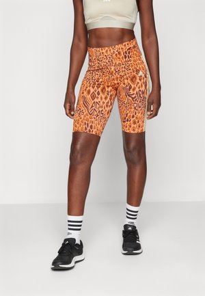 Atleet die oranje gebloemde Adidas fietsbroek draagt, een beige sportbeha, zwarte sneakers en witte Adidas sokken, staand tegen een effen achtergrond.