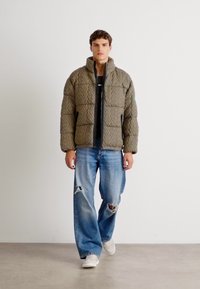 Quilted olivengrøn pufferjakke med høj krave, lynlås lukning og skrå lommer; båret over en sort skjorte og slidte lys blå jeans.