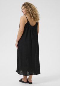 Vestido maxi negro con un diseño fluido, tirantes ajustables y un escote con volantes. La parte trasera cuenta con un detalle de lazo. Se lleva con sandalias negras.