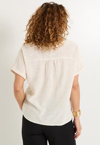 Blusa corta de mangas con un tono beige claro, con un tejido texturizado, que presenta un detalle de pliegue en la parte posterior y puños doblados, combinada con pantalones negros.