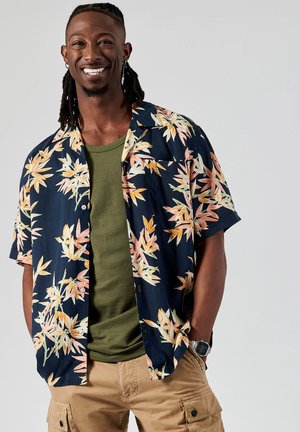Chemise à manches courtes bleu marine avec un motif floral orange et crème, superposée sur un T-shirt vert, assortie avec un short cargo beige.