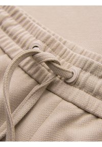 Beige textured fabric sweatpants with elastic waistband and tied drawstring threaded through metal eyelets.