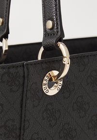 Sac à main noir texturé avec quincaillerie dorée, présentant un charme circulaire portant le nom de la marque et de solides poignées en cuir.