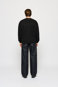 Man standing facing away wearing a black long-sleeve shirt with "eightyfive" text on back and loose dark blue jeans.