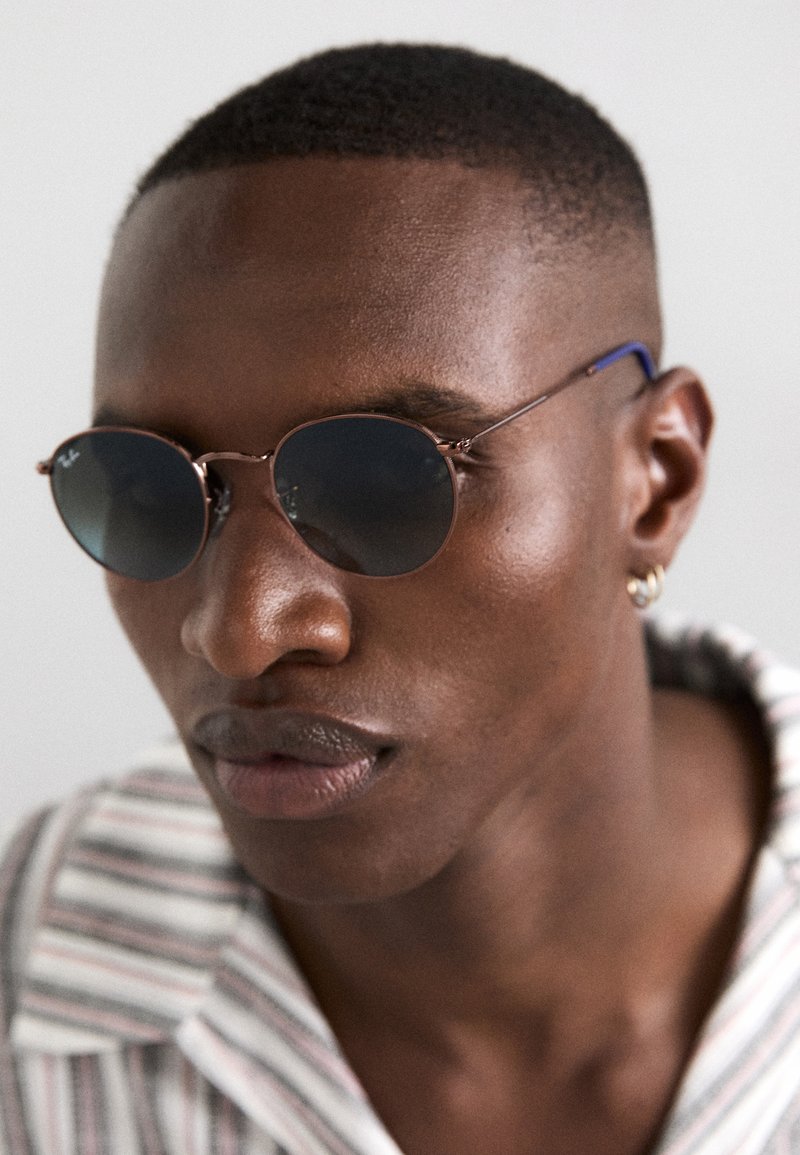 Round sunglasses with dark lenses, thin metal frame in rose gold, featuring blue accents on the temple arms. Smooth skin texture, neutral backdrop.