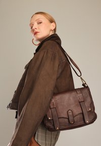 Brun læder crossbody-taske med to frontremme, messinghardware og en tekstureret finish. Båret af en model i en brun jakke.