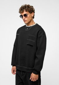 Zwarte sweatshirt van zacht materiaal, met een borstzak, geribbelde mouwen en contrasterende witte stiksels. Draag het met een zonnebril.