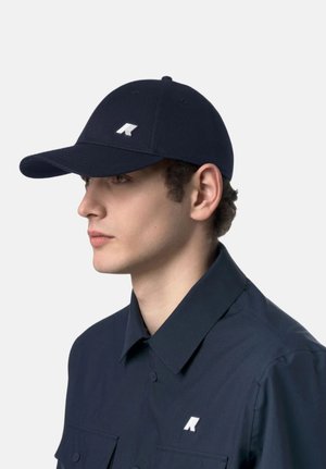 Giovane uomo con indosso un cappellino blu navy e una camicia coordinata con bottoni, entrambe con un logo geometrico bianco sul lato sinistro.