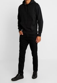 Schwarzer Hoodie mit Fronttasche, Kapuze mit Kordelzug und gerippten Bündchen. Kombiniert mit schwarzen Slim-Fit-Jeans und schwarzen Schnürschuhen.