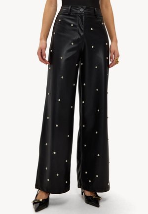 Pantalones de cuero sintético negro de pierna ancha con tachuelas metálicas dispersas, combinados con tacones negros de punta afilada y una blusa negra.