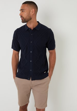 Marineblauwe cardigan met korte mouwen, een gestructureerd ruitpatroon, knoopsluiting aan de voorkant en ribgebreide onderrand. Gecombineerd met beige shorts.