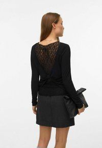 Žena u crnoj bluzici dugih rukava s čipkastim detaljem na leđima i tamnosivoj suknji, drži crnu presavijenu torbicu.