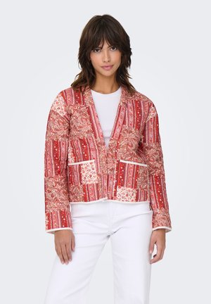 Femme portant une veste matelassée rouge à motifs par-dessus un haut blanc et un pantalon blanc, debout devant un fond clair uni.