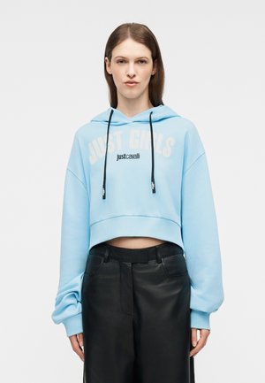 Tânără purtând un hanorac scurt, albastru deschis, cu textul "JUST GIRLS" și "justcavalli", asortat cu pantaloni negri din piele, cu talie înaltă.