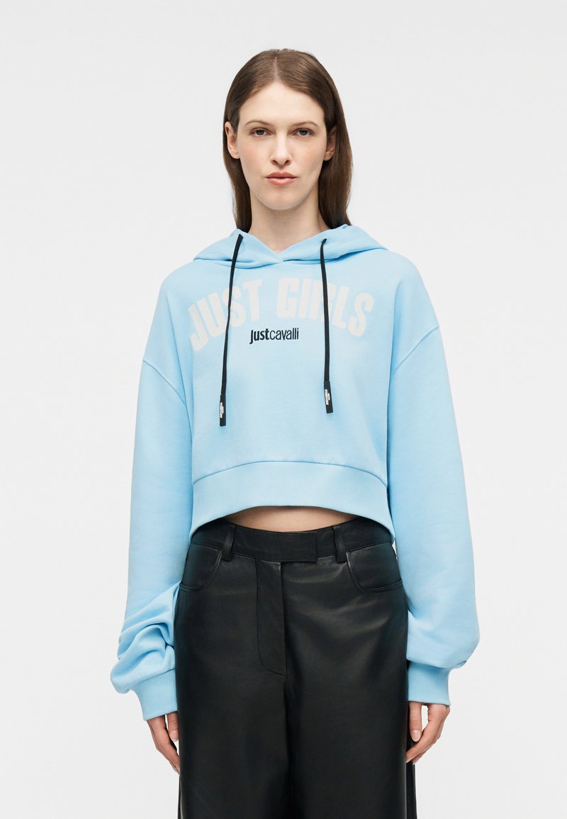 Νεαρή γυναίκα που φοράει γαλάζιο cropped φούτερ με τα κείμενα «JUST GIRLS» και «justcavalli», συνδυασμένο με μαύρο δερμάτινο παντελόνι ψηλόμεσο.