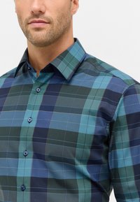 Uomo che indossa una camicia a quadri blu, verde e nera con colletto, bottoni e parte della sua barba e mascella visibili.
