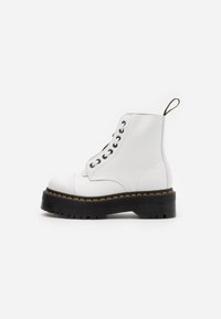 Botas de tobillo de cuero blanco con una gruesa suela de goma negra, que presentan un diseño de cordones y costuras amarillas a lo largo del borde de la suela.