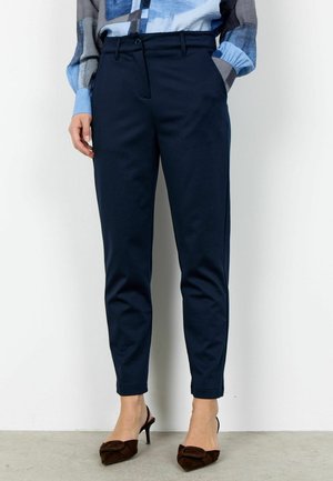 Nő sötétkék, szűk szárú nadrágban, kék mintás blúzban, amelynek felhajtott az ujja, és barna, alacsony sarkú, hegyes orrú cipőben, fehér fal előtt áll.