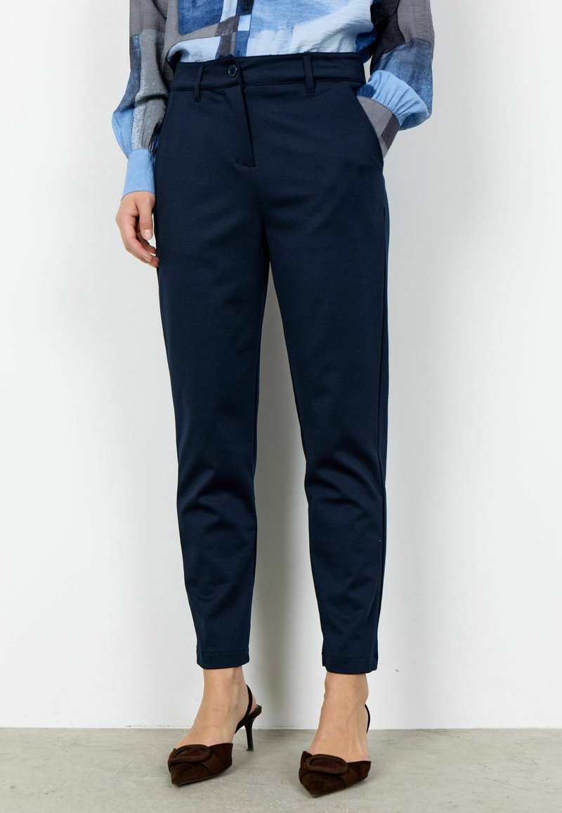 Žena oblečená v tmavomodrých zúžených nohaviciach, modrej vzorovanej blúzke s vyhrnutými rukávmi a hnedých nízkom podpätkoch s ostrou špičkou stojí opretá o bielu stenu.