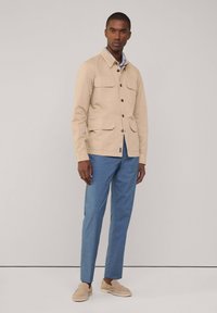 Beige Baumwolljacke mit vier vorderen Taschen, Knopfverschluss und Hemdkragen, kombiniert mit hellblauen maßgeschneiderten Hosen und beigen Slippers.