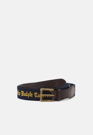 Granatowy pasek materiałowy z żółtym napisem „Polo Ralph Lauren”, ciemnobrązowymi skórzanymi końcówkami i mosiężną sprzączką z podwójnym ząbkiem na białym tle.