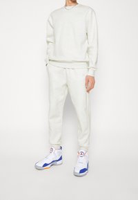 Person klädd i en ljusbeige matchande sweatshirt och sweatpants med vita, blå och oranga högskurna sneakers, stående framför en enkel bakgrund.