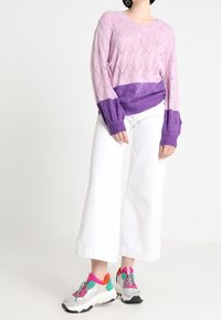 V-ringad, stickad tröja i rosa och lila med diamantmönster; ihop med vita vida byxor och färgglada klumpiga sneakers.
