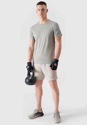 Młody mężczyzna w szarej sportowej koszulce i szortach, czarne bezpalcowe rękawiczki, trzyma kettlebell o wadze 8 kg, stoi na jednolitym jasnym tle.
