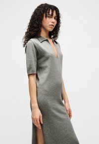 Γυναίκα με σγουρά μαλλιά, φοράει γκρι μάλλινο φόρεμα με γιακά, βαθύ V λαιμόκοψη, μισά μανίκια και σκίσιμο στο πλάι, στέκεται μπροστά σε απλό φόντο.