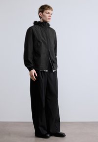 Must kerge jope kapuutsiga, lukuga ja elastsete varrukaservadega, koos lahtiste mustade pükste ja mustade jalanõudega.