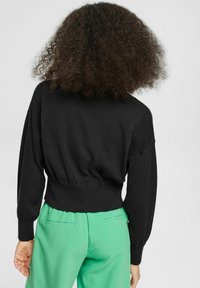 Maglione nero a pullover con texture a coste, maniche a palloncino e vita aderente, indossato sopra pantaloni verdi.